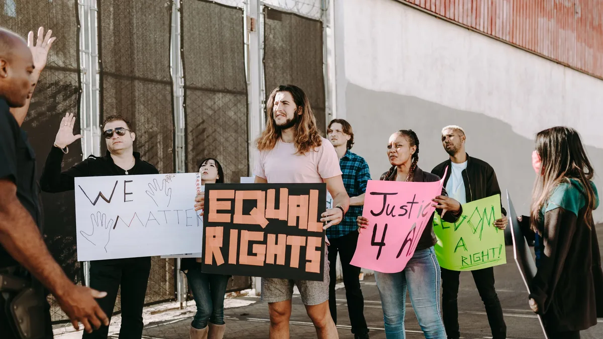 A group of people are gathered outside, holding signs that read "EQUAL RIGHTS" and "Just 4 A RIGHT!" The individuals are dressed in casual attire, with some wearing jeans and others in more formal clothing. The scene appears to be a protest or demonstration, with the group standing together in solidarity.